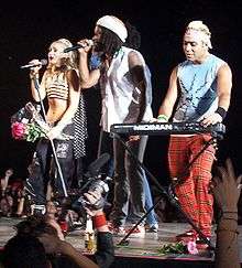 A blonde lady holding a bunch of pink flowers (Stefani), a black man with dreadlocks (McNair) and an Indian man with dyed-blond hair (Kanal) stand on a grey stage. The first two performers are singing into microphones, while the third performer plays a keyboard. In the background, a crowd cheers them. In the foreground, a man holds up a camcorder to film the group.
