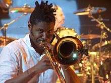 Gabrial McNair—a black male with dreadlocks, wearing a button-up pinstripe white shirt—plays trombone