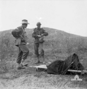 A soldier lies on a stretcher covered by a blanket, while two Caucasian men in uniform stand above him smoking cigarettes