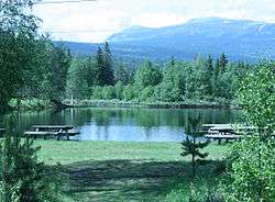 A landscape showing a grassy area with park benches near a lake, surrounded by forest and mountains