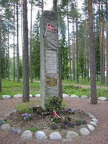 A stone monument in a forest clearing, adorned with a plaque and small Norwegian flag