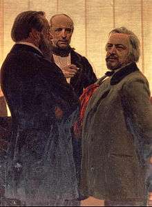Three men standing together – two men with beards, the one on the right with grey hair, flanking a third man watching them intently