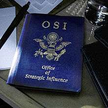 A blue book similar to an American passport lies on a desk surrounded by papers, folders, a pen and a glass of water. The blue book has "OSI" printed on its top centre, and "Office of Strategic Influence" along the bottom centre of the book in smaller text. The eagle found on American passports has been altered: the arrows are double-headed, the olive branch is wilted, and the eagle's wings have been clipped.