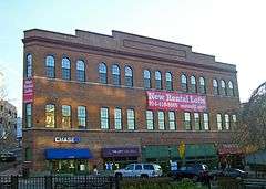 A large three-story brick building with storefronts on the bottom, rounded-arch windows on the second and third levels and a parapet on top. Hanging from the front is a banner with "New rental lofts" and a phone number.