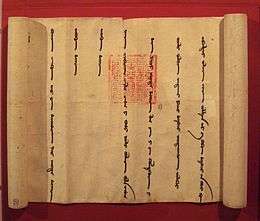 A partially unrolled scroll. opened from left to right to show a portion of the scroll with widely spaced vertical lines of cursive Mongol script. Imprinted over two of the lines is an official-looking square red stamp with an intricate design.