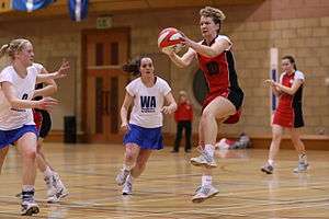 Netball players in centre third of court. One player in red has caught the ball and two players in white are moving towards her.