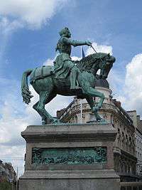 Photograph of a bronze statue of Joan of Arc on a horse, all on a large plinth. The picture is taken from below. She is in armour and carries a sword in her outstretched right hand. The horse has its head bowed and its right foreleg raised, as if trotting. The bronze statue is green from verdigris.