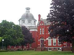 Two red buildings, both slightly obscured by trees in front of them. The one on the left has a metallic dome, the one on the right has a pointed roof with chimney