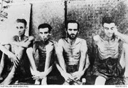 Four malnourished shirtless men sit against a wall