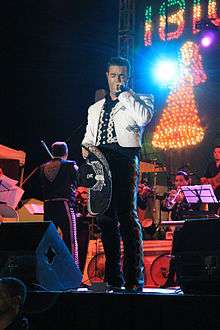 A man dressed in Mexican charro suit in black and white, holding a microphone in his right hand and a hat on the left.