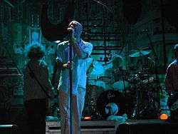 A blue-tinted photograph of musicians performing in front of an industrial background. From left to right: a long-haired male stands with his back to the camera playing bass guitar, a middle-aged Caucasian male sings into a microphone, a middle-aged Caucasian male plays behind a black-and-silver drum set on a riser, and a guitar player is mostly cropped from the extreme left of the photo.