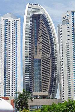 A tall, white- and grey-colored building with a curved facade, towering above shorter buildings nearby. This is the Trump Ocean Club International Hotel and Tower in Panama City, Panama