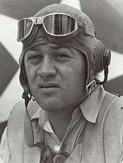 Head of a squinting man wearing a shirt unbottoned at the collar and a cloth aviator's cap with headphones built into the ear flaps, an unbuckled chin strap, and goggles pushed up onto his forehead.