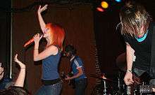 A band performs for a live audience. Two men are playing guitars while a woman with bright red hair sings into a microphone.