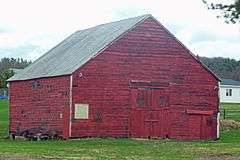 A red wooden barn with a pointed roof. Some buildings and a low wooded ridgeline are at the rear.