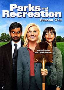 The DVD cover shows three people standing side-by-side. In the middle, a blond woman wears a gray dress suit, smiling and holding a golden shovel. On the right, a brown-haired woman in a green jacket looks at her and smiles. On the left, a black-haired man with a beard, wearing a gray suit and green tie, looks at the middle figure while smirking. Above the trio is the text, "Parks and Recreation Season One".