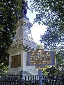 A tall stone pedestal with a metallic figure atop it, with two large trees above it. The base is surrounded by an iron fence. In the front right is a blue marker with yellow type headed "André Captured", over smaller type describing that event.