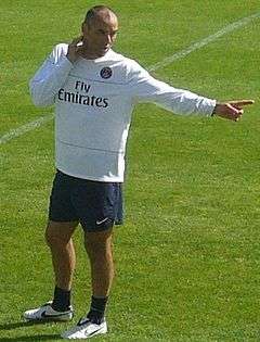 A middle-aged man wearing a white shirt, black shorts and white trainers, standing on a grass field.