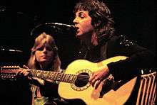McCartney, seated, playing a twelve-string acoustic guitar, Linda McCartney can be seen seated to his right.