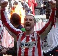 A man wearing a red and white football shirt after a game; with his arms raised triumphantly.