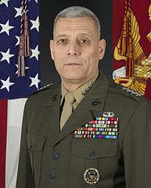 A color image of John Paxton, a white male in his Marine Corps Service A uniform. He is not wearing a hat, ribbons are visible as well as a basic parachutist badge. The Marine Corps flag and United States flag are visible in the background.