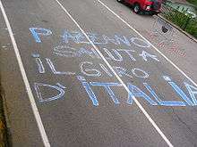 Overhead view of a road with words in Italian written on it in blue chalk. A red vehicle is partly visible on the right side of the street, as is a black fence and one section of a steel guardrail, with red and white rope on it.