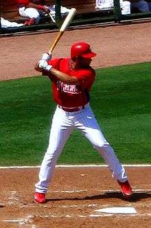 A right-handed man in a red batting practice baseball jersey and white baseball pants with red pinstripes stands in a batter's box by home plate.