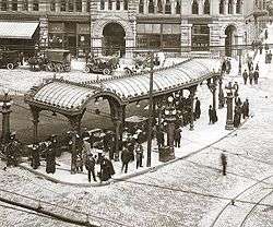 Historic photograph of the Pioneer Square Pergola with the Pioneer Building behind. Many people wait under the Pergola as the cobbled streets bustle around them.