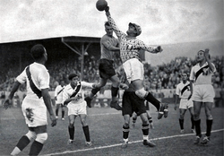 An action shot from a football match. A goalkeeper jumps and punches the ball away from his goalmouth