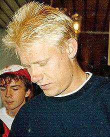 Peter Schmeichel, with blonde hair and wearing a dark blue sweater, looks down towards his left with an unidentified man in the left background.