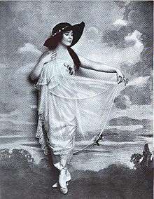 Black and white full-length portrait of a white woman wearing a white dress and a dark hat.
