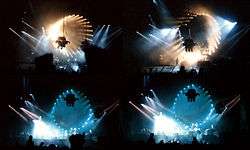 Four images, arranged to form a rectangle, of a darkened concert venue. The stage is dominated by a circular pattern of lights which surround a projection screen. Members of the audience are silhouetted in the foreground.