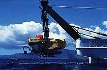 A submarine, hanging from the back of a ship via a crane, is lowered into the water.