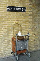 A sign reading "Platform 9¾" with half of a luggage trolley installed beneath, at the interior of King's Cross railway station.