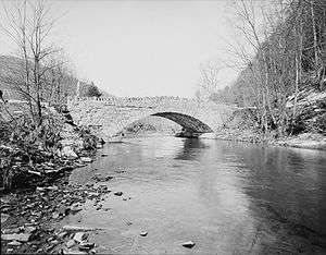 Black and white photograph of a cracked stone bridge partly reflected in a stream. Both of the stream banks are wooded.