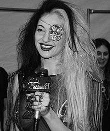 A woman with long hair, half of it being blonde and the another half being dark wearing an eyepatch in her left eye talks to a microphone.