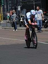 A road racing cyclist wearing a light blue skinsuit and matching aerodynamic helmet, with pink shoes and gloves. Spectators watch at the roadside.