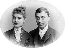  A charming and iconic period photo of a young couple around 1900. The lady has her hair swept upwards and is wearing a high collar fastened with a jewel. The gentleman sports a gallant moustache neatly trimmed and is also wearing a high collar.
