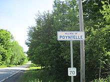 An image of a white sign with blue lettering and border, reading "VILLAGE OF/POYNTELLE," against a background of greenery.