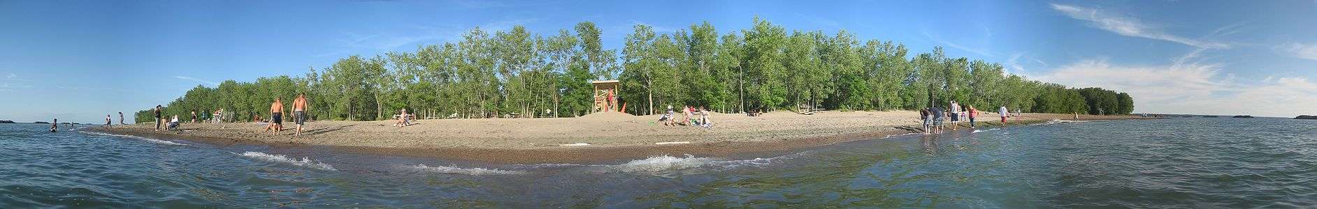 A view of a sunlit beach, that is flanked by green trees, from the water under a mostly clear, blue sky with several vacationers.