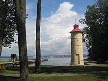 A small off-white lighthouse with a red roof and the trunks of two trees overlooking a bay with a city in the background.