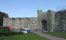 Quebec's restored city wall is gray stone about 20 feet (6.5 meters) high.  The St. John's gate has a modern road going through it, and has a copper-roofed turret on the left bastion.  A paved path goes through a grassy area below the wall.