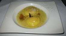 A pair of ravioli are immersed in egg yolk and topped with shredded cheese. The dish is served on a white, rectangular dish with a bowl shape in the center.