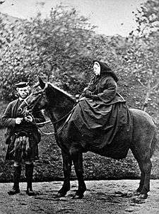 A woman, severely  dressed in black, seated on a horse with a man standing by its head.