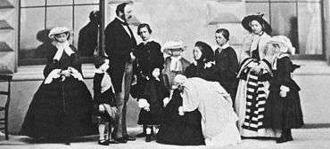 Photograph of a seated Victoria, dressed in black, holding an infant with her children and Prince Albert standing around her