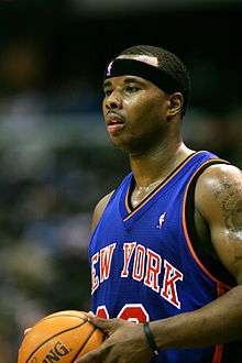 Quentin Richardson is profiled looking to the left while he is holding a basketball. He is wearing a blue New York Knicks uniform.