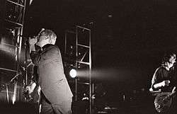 A black-and-white photograph of Michael Stipe and Peter Buck performing on stage with spotlights on them. Stipe is to the left singing into a microphone, wearing a three-piece suit, he has bleach-blond hair and is obscuring Mike Mills, whose bass guitar is visible from behind him. Peter Buck is playing guitar and wearing a button-up pattern shirt behind Stipe to the photograph's right with a sneer on his face.