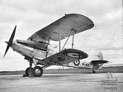 Photograph of a biplane on a cement tarmac. The image is taken from a lower angle on the side of the aircraft. The identification number A1-62 can be seen on the tail, beside a symbol of an outlined circle with an enclosed circle in its centre (the RAAF roundel).