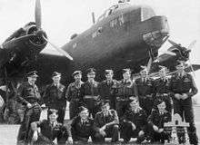 Black-and-white photo of a group of young men wearing military uniform in a formal pose in front of a larger World War II-era propeller aircraft
