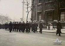 Black-and-white photo of a large group of men wearing military uniforms marching in formation along a city street